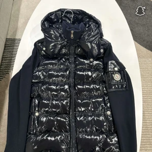 Svart dunväst från Moncler med huva - Säljer en svart dunväst från Moncler med glansig yta och avtagbar huva. Västen har ribbade stickade partier på sidor och ärmar, dragkedja framtill och Moncler-logga på ärmen. Insidan har en cool serietidningsetikett. Perfekt för dig som gillar exklusiva streetwear-vibes.