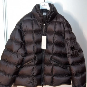 C.P. Company jacka  - Snygg svart dunjacka från C.P. Company med puffad look och hög krage. Jackan har dragkedja framtill, två snedställda fickor med dragkedja och den ikoniska linsen på ärmen. Perfekt för kalla dagar och riktigt clean stil. Köptes för 7600kr på jonhells endast testad kommer med extra knappar tags Size 54 passar L/xl