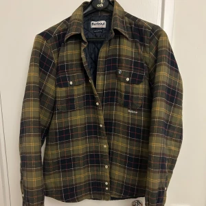  Barbour Overshirt  - Snygg rutig overshirt från Barbour i gröna, gula och röda toner. Skjortan har två bröstfickor med knappar och klassisk krage. Perfekt lager-på-lager-plagg med quiltat innerfoder och knäppning framtill.