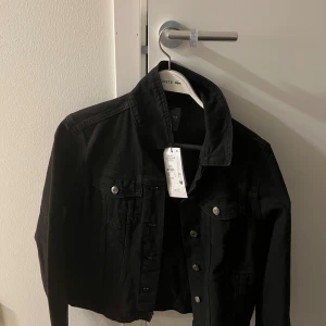 Svart jeansjacka från Primark - Säljer en svart jeansjacka från Primark i klassisk modell med knappar framtill och två bröstfickor. Jackan är tillverkad av bomull med inslag av återvunnen bomull och har en snygg, tidlös look som passar till det mesta.