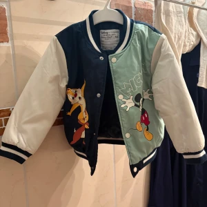 Disney bomberjacka med Mickey & Piff och Puff - Snygg bomberjacka från Disney x Primark med stora tryck av Mickey Mouse samt Piff och Puff. Jackan har vita ärmar, ena sidan är marinblå och andra ljusgrön, med ribbade muddar och tryckknappar framtill. Perfekt för dig som älskar Disney och vill sticka ut.