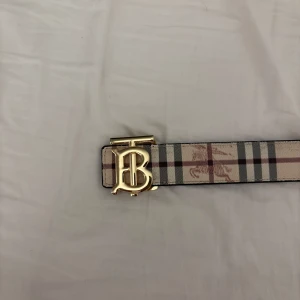 Burberry rutigt bälte med guldspänne - Säljer ett snyggt Burberry-bälte med klassiskt rutigt mönster i beige, svart, grått och rött. Bältet har ett stort guldigt spänne med Burberrys ikoniska monogram. Tillverkat i skinn och perfekt för att lyfta vilken outfit som helst.