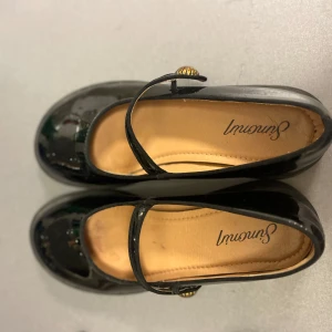 Svarta lackade ballerinaskor  - Stilrena svarta ballerinaskor i glansigt lackmaterial. Skorna har en klassisk Mary Jane-rem med snygg guldfärgad knappdetalj och rund tå. Perfekta för dig som gillar en söt och tidlös look med en twist.