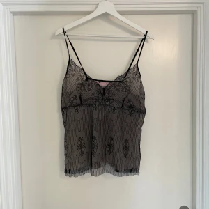 Svart spetsig linnetopp från hunkemöller - Säljer en svart, transparent linnetopp från hunkemöller med tunna axelband och vacker spetsdetalj. Toppen har ett unikt mönster med broderade ornament och subtila ränder, samt en lätt v-ringad hals. Perfekt för dig som gillar en edgy och feminin stil.