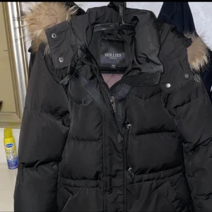 Svart pufferjacka med päls från Hollies - Säljer en svart pufferjacka från Hollies med fluffig päls på huvan. Jackan har dragkedja och knappar framtill, samt en snygg, quiltad design. Perfekt för kalla vinterdagar och har en riktigt skön oversized känsla. Huvan är stor och pälsen är ljusbrun.