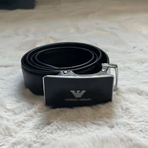 Svart skärp från Giorgio Armani - Stilrent svart bälte från Giorgio Armani med en bred, blank silverfärgad spänne och logga framtill. Bältet är gjort i slätt skinn och passar perfekt till att lyfta vilken outfit som helst med en lyxig touch.