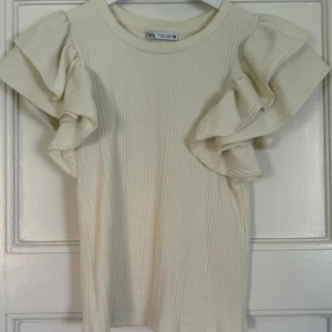 Ribbad volangtop från Zara - Superfin ribbad topp från Zara i ljus beige med volangdetaljer på axlarna. Toppen har rund halsringning och är kortärmad, perfekt för att lyfta vilken outfit som helst med sina snygga volanger.