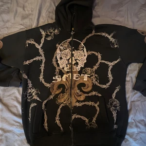 Svart hoodie med dödskalle från Smet - Svart hoodie från Smet med dragkedja, huva och feta tryck av dödskallar, taggtråd och rosor både fram och bak. Texten 'Born on the street Los Angeles' på ryggen. Grym streetstyle med detaljer som sticker ut.