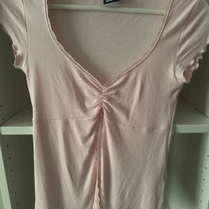 Ljusrosa topp med slit och spets - Ljusrosa topp från Gina Tricot med slit och v-ringning. Toppen har korta ärmar, spetskant längs urringningen och framtill samt snygg rynkdetalj i mitten. Säljs inte längre men är i storlek xs och har används två gånger💞