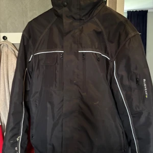 Svart vindjacka från Bluewear XL - Svart vindjacka från Bluewear i storlek XL. Jackan har flera dragkedjor, praktiska fickor och vita kontrastsömmar. Perfekt för blåsiga dagar och har en sportig look med hög krage och detaljer på ärmarna. Den har några fläckar eftersom den använts i arbete