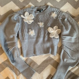 Ljusblå stickad tröja med blommor - Superfin ljusblå stickad tröja från SHEIN MOD med puffiga långärmar och coolt vågigt mönster på bröstet. Dekorerad med stora vita 3D-blommor som ger en unik vibe. Perfekt för dig som vill sticka ut med en söt och trendig look.
