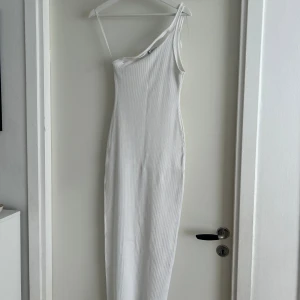 Vit ribbad långklänning oneshoulder - Säljer en vit ribbad långklänning med oneshoulder och smal axelrem. Klänningen har en slits på sidan och är figurnära med stretchigt material. Perfekt för dig som gillar minimalistisk och stilren design.