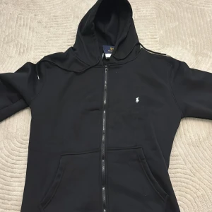 Svart hoodie från Polo Ralph Lauren - Svart hoodie från Polo Ralph Lauren med klassisk vit broderad logga på bröstet. Tröjan har huva med snörning, dragkedja framtill och två stora fickor. Materialet är mjuk bomull och passformen är normal. Perfekt för chill dagar och streetwear.