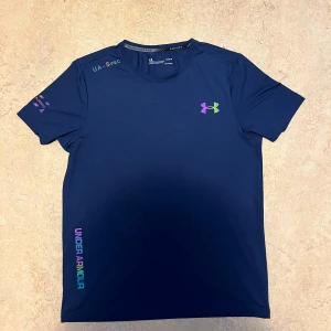 Blå Under Armour t-shirt - Snygg marinblå t-shirt från Under Armour med korta ärmar och rund hals. Loggan och texten på bröstet och sidan har coola färger i lila, grön och blå. Perfekt för träning och sport, tillverkad i lätt polyester som andas.