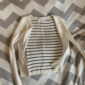 Vit och svart randig kofta från C'est Monique - Säljer en ribbad kofta från C'est Monique i vit med svarta horisontella ränder. Koftan har lång ärm, rund hals och knappar hela vägen framtill. Materialet är mjukt och stretchigt, perfekt för lager-på-lager. Passar dig som gillar stilrena och klassiska plagg.