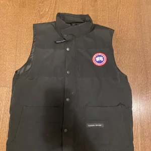 Svart väst från Canada Goose - Svart dunväst från Canada Goose med klassisk logga på bröstet och hög krage. Västen har två stora fickor framtill och stängs med både dragkedja och tryckknappar. Perfekt för lager-på-lager och riktigt snygg till streetwear.