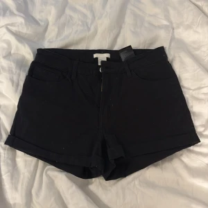 Svarta shorts från H&M, strl 36 - Svarta shorts från H&M i klassisk femficksmodell med uppvikta benslut och dragkedja framtill. Snygg midwaist-passform och tillverkade i mjuk bomull som är skön mot huden. Perfekta för varma dagar och enkla att matcha med din favorit-topp.