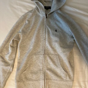 Grå hoodie från Polo Ralph Lauren - Snygg ljusgrå hoodie från Polo Ralph Lauren med dragkedja och klassisk broderad logga på bröstet. Tröjan har huva, kängurufickor och är tillverkad i mjuk bomullsmix. Perfekt för chill dagar eller när du vill ha en clean och stilren look.