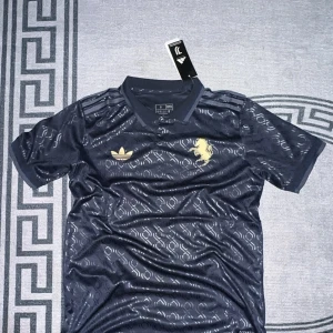 Juventus x Adidas marinblå fotbollströja - Äkta Juventus x Adidas fotbollströja i marinblått med guldiga detaljer. Tröjan har korta ärmar, krage och ett snyggt geometriskt mönster över hela tyget. Juventus-logga i guld på ryggen och Adidas Trefoil samt Juventus-lejon på bröstet. Tillverkad i lätt, glansigt material.