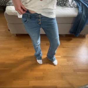 Blå bootcut jeans med knappar - Snygga blå jeans med bootcut-modell och flera synliga knappar i gylfen. Jeansen har klassisk femficksdesign och är tillverkade i ett mjukt denimtyg. Perfekta för dig som gillar en lite retro vibe och vill ha ett par jeans som sticker ut.