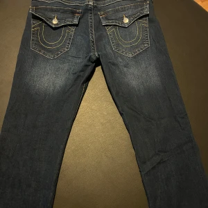 True Religion Ricky jeans mörkblå - Mörkblå jeans från True Religion, modell Ricky. Klassiska kontrastsömmar i gult och orange, stora fickor med lock och dekorativa sömmar bak. Grova detaljer och logopatch bak i midjan. Snygg passform och cool streetstil.