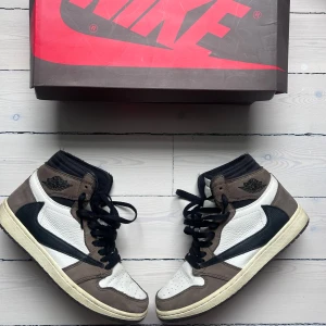 Nike Air Jordan 1 Travis Scott High - Nike Air Jordan 1 Travis Scott High med ikonisk omvänd Swoosh i svart, bruna mockadetaljer och vitt läder. Höga sneakers med svarta snören och klassisk Air Jordan-logga på sidan. Perfekt för dig som vill sticka ut med en unik och eftertraktad sneaker. Tillkommer med extra laces och box, har fysiskt kvitto. Öppen för bud, endast seriösa. 