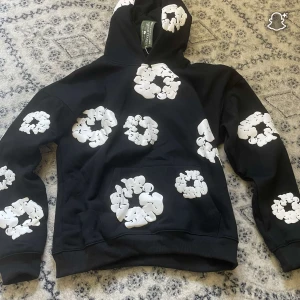 Svart hoodie från Denim Tears med blommor - Svart hoodie från Denim Tears med stora vita blommönster över hela tröjan, inklusive huva och ärmar. Tröjan har huva och magficka, och är tillverkad i mjuk bomull. Perfekt för dig som vill sticka ut med en unik och trendig look.