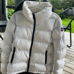Vit pufferjacka från Colmar - Säljer en snygg vit pufferjacka från Colmar med svart dragkedja och två fickor framtill. Jackan har hög krage och klassisk quiltad design, perfekt för kalla dagar. Märkeslogga på ärmen och riktigt skön passform. Stl 44, passar dig som vill ha en stilren vinterlook.
