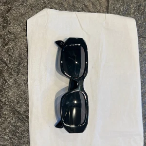 Svarta solglasögon från Prada - Säljer ett par svarta solglasögon från Prada med chunky, rektangulär form och breda skalmar. Glasögonen har en cool, modern vibe och är tillverkade i blankt syntetmaterial. Prada-loggan syns tydligt på sidan av skalmarna. Perfekt för dig som vill sticka ut.