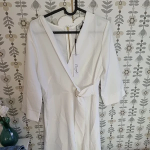 Vit omlottklänning från Closet - Snygg vit omlottklänning från Closet med trekvartsärm och v-ringad hals. Klänningen har ett elegant bälte med guldfärgad ring i midjan och stilren design. Perfekt för dig som gillar minimalistisk och chic stil.