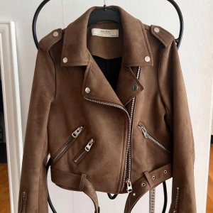 Brun mocka bikerjacka från Zara - Snygg brun bikerjacka i mockaimitation från Zara Basic. Jackan har coola dragkedjedetaljer, bred krage och bälte i midjan. Perfekt för dig som vill ha en edgy look med stilrena vibbar. Passar till allt från jeans till klänning.