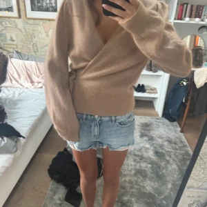 Beige omlottkofta i kashmir - Supermjuk beige omlottkofta i kashmir med knyt i sidan och v-ringad hals. Långärmad och med en loose fit som ger en chill och trendig vibe. Perfekt att slänga på sig över ett linne eller t-shirt för en snygg lager-på-lager-look.