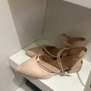 Beiga pumps med spetsig tå och rem - Snygga beiga pumps med spetsig tå och låg klack. Skorna har en elegant rem med glittriga detaljer och justerbart spänne runt ankeln. Perfekta för dig som vill ha en stilren och trendig look.