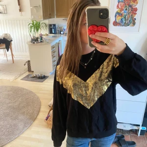 Svart sweatshirt med guld V-detalj - Cool svart sweatshirt med en stor glansig V-formad detalj i guld framtill. Tröjan har rund hals och långa ärmar med muddar. Perfekt för dig som vill sticka ut lite extra med metallic vibes.