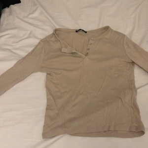 Beige långärmad topp från Zara - Säljer en beige långärmad topp från Zara med knappar vid halsringningen. Tunt och mjukt material som känns skönt mot huden. Perfekt att styla med jeans eller kjol för en chill look.