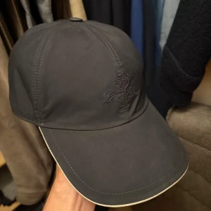 Loro Piana keps  - Snygg navy keps från Loro Piana med broderad logga framtill. Utsidan är slät och stilren, insidan har ett rutigt mönster i beige, rött och grönt. Skärmen är beige undertill och kepsen har elastiskt band baktill för skön passform. Perfekt för dig som gillar exklusiva accessoarer.