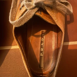 Mocka skor - Snygga bruna sneakers i mjuk mocka med rund tå och platt sula. Skorna har breda snören och diskret Prada-logga på sidan och innersulan. Perfekta för dig som gillar stilrena och exklusiva sneakers med en klassisk vibe. Ni ser vad de är för märke! 