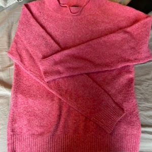 Rosa stickad tröja med hög krage - Mysig rosa stickad tröja med hög krage och långa ärmar. Tröjan har breda ribbade muddar vid ärmslut och nederkant, perfekt för kyliga dagar. Enkel och stilren design som är lätt att matcha med dina favoritjeans eller kjol.