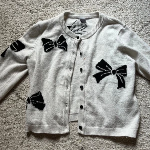 Vit kofta med svarta rosetter från Lindex - Vit kofta från Lindex med svarta rosetter som mönster. Rund hals, svarta knappar framtill och mjukt stickat material. 