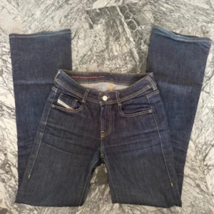 Mörkblå jeans från Diesel, stl 24/30 - Snygga mörkblå jeans från Diesel med klassisk femficksdesign och kontrastsömmar. Modellen är rak och har en normal passform. Jeansen är tillverkade i slitstarkt denim och har en cool läderpatch bak med Diesel Industry-logga.