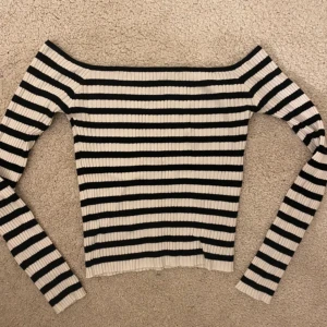Randig offshoulder topp House Brand - Säljer en ribbad offshoulder topp från House Brand i storlek M. Toppen är svart och vit med horisontella ränder och har långa ärmar. Den är figurnära och har en snygg, croppad passform som passar perfekt till jeans eller kjol.