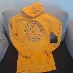 Gul Gymshark hoodie med tryck S - Gul hoodie från Gymshark i storlek S med blått tryck av en muskulös haj och texten 'GYMSHARK FITNESS' på bröstet. Tröjan har huva med snörning, känguruficka och långa ärmar. Perfekt för gymmet eller chill dagar.