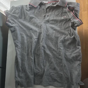 Grå pikétröja från Moncler - Snygg grå pikétröja från Moncler med klassisk krage och knäppning framtill. Tröjan har korta ärmar med röd och vit randig detalj på krage och ärmslut samt Moncler-logga på bröstet. Perfekt för en stilren och sportig look.