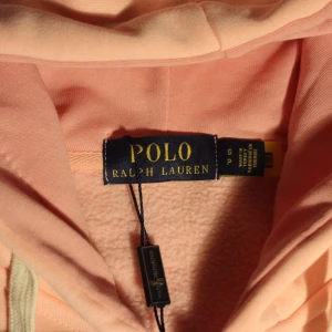 Ljusrosa hoodie från Polo Ralph Lauren - Säljer en ljusrosa hoodie från Polo Ralph Lauren med dragkedja och huva. Tröjan har vita snören, två fickor fram och den klassiska broderade loggan i mörkblått på bröstet. Perfekt för dig som gillar stilrena och bekväma plagg. Den är helt ny och aldrig använd