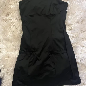Svart axelbandslös kort klänning - Snygg svart axelbandslös klänning med figurnära passform och stilren design. Klänningen är kort och har diskreta fickdetaljer framtill.  Aldrig använd