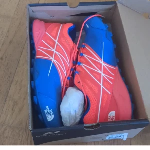 Blå och röda fotbollsskor The North Face - Snygga fotbollsskor från The North Face i blått och rött med vita detaljer och snörning. Skorna har mönstrad ovandel och dobbar för bra grepp på planen. Perfekta för dig som vill sticka ut med färg och stil på matchen.