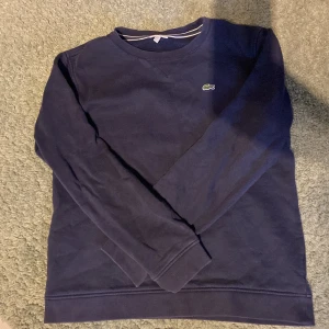 Mörkblå sweatshirt från Lacoste  - Mörkblå sweatshirt från Lacoste  med klassisk rund hals och broderad krokodil-logga på bröstet. Tröjan har långa ärmar och är tillverkad i mjuk bomull, perfekt för en chill och sportig stil. Lite solblekt men ingenting man märker av. Hör av er vid minsta funderingar!