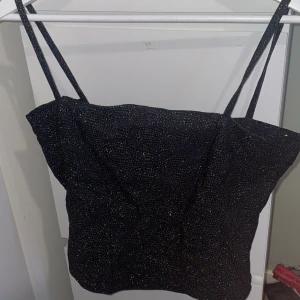 Glittrig svart festtopp - Snygg svart linne topp med tunna axelband och glittrigt material som fångar ljuset. Perfekt till fest, nyår eller utekväll. Storlek XL men kan upplevas lite tajt så passar även L.