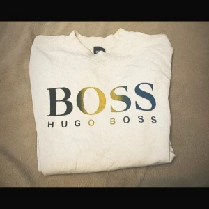 Vit sweatshirt från Hugo Boss - Vit sweatshirt från Hugo Boss med stor BOSS-logga i svart, gult och blått framtill. Tröjan har rund hals, långa ärmar och ribbade muddar vid ärmslut och nederkant. Perfekt för en clean och stilren look.