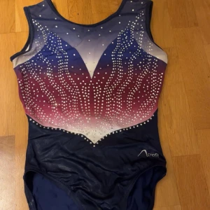 Glittrig gymnastikdräkt  från veraspory - Snygg gymnastikdräkt från Vera sport i blått, lila och vitt med glittriga stenar framtill. Dräkten har ärmlös design och en tight passform, perfekt för träning eller tävling. Materialet är syntet och ger en glansig finish.